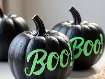 Two black pumpkins with the word “BOO!” written in bright green letters sit on a white surface, with another pumpkin and some beige straw partially visible in the background.