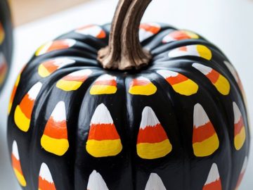 A pumpkin painted black with candy corn designs in yellow, orange, and white covering its surface, displayed on a light-colored surface.