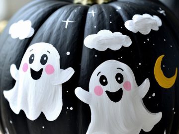 A black pumpkin painted with two smiling white cartoon ghosts, white clouds, stars, and a yellow crescent moon, creating a cute Halloween-themed design.