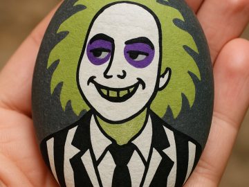 A hand holds a smooth stone painted with a cartoon version of Beetlejuice, featuring green hair, pale skin, purple eyeshadow, a wide grin, and a black-and-white striped suit.