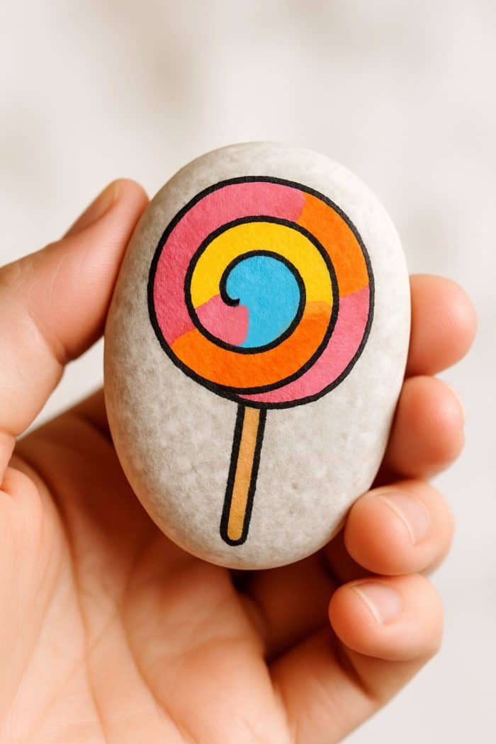 A hand holding a smooth white stone with a colorful lollipop design, featuring pink, yellow, orange, and blue swirls, painted on its surface.