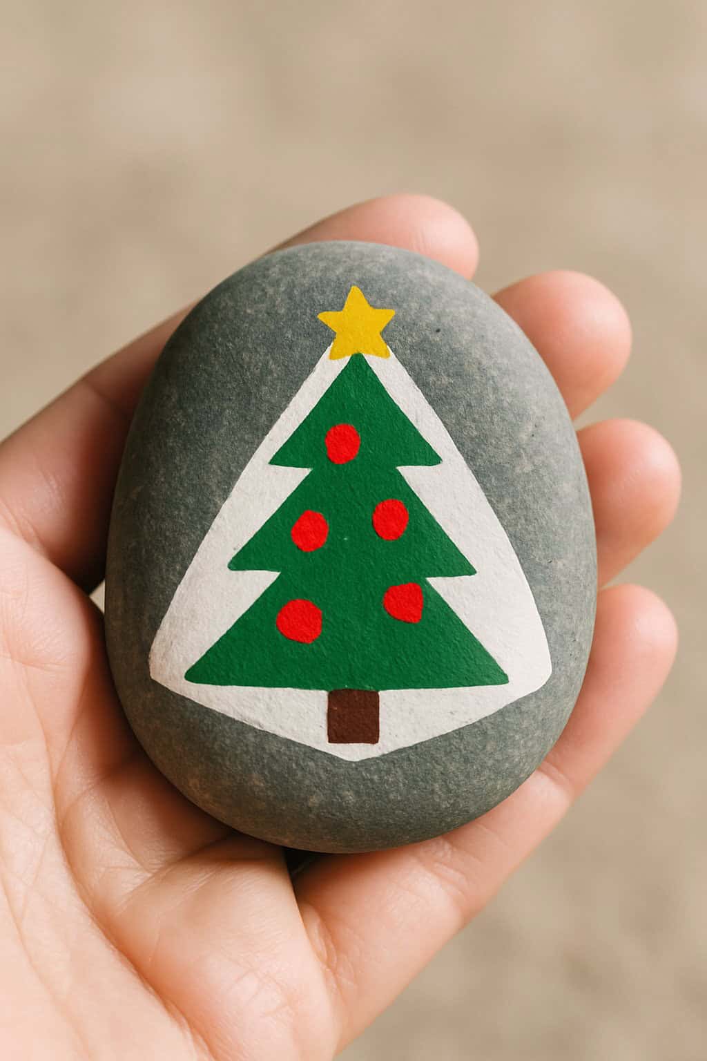 A hand holds a smooth, oval stone painted with a green Christmas tree decorated with red ornaments, a yellow star on top, and a brown trunk.
