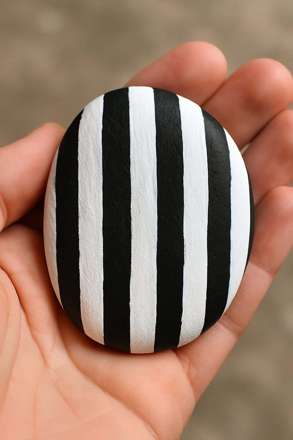 A hand holding a smooth river rock painted with black and white vertical stripes.