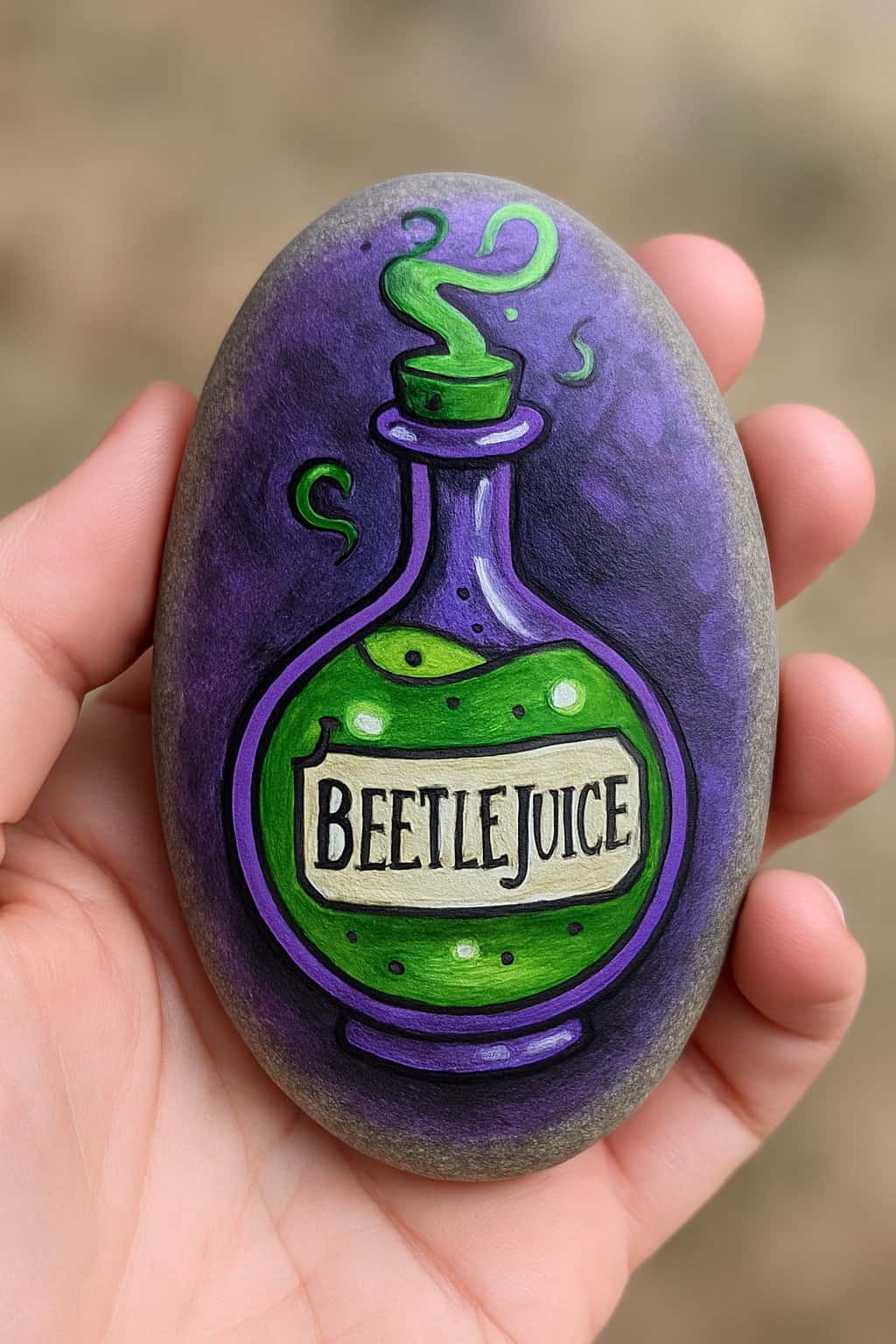 A hand holding a smooth painted rock with a colorful beetlejuice potion bottle design.