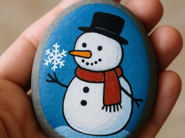 A hand holds a smooth stone painted with a cheerful snowman wearing a black hat and red scarf, with a snowflake near its stick arm, set against a blue background.