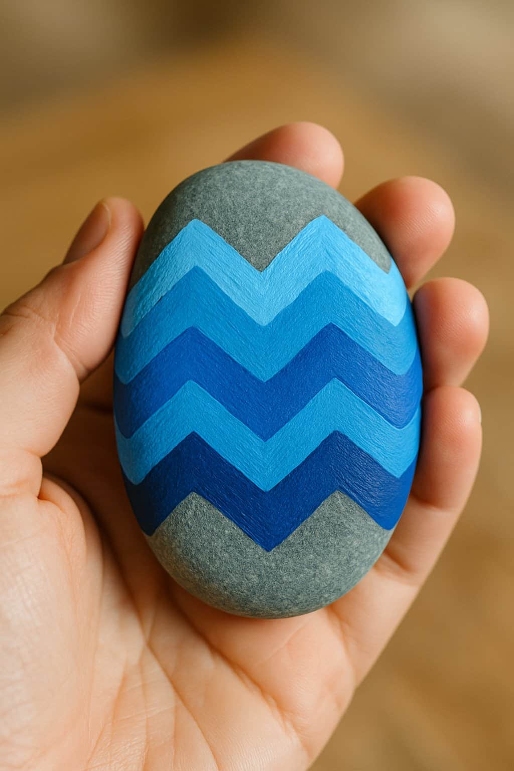 A hand holding a smooth river rock painted with a blue zigzag pattern.