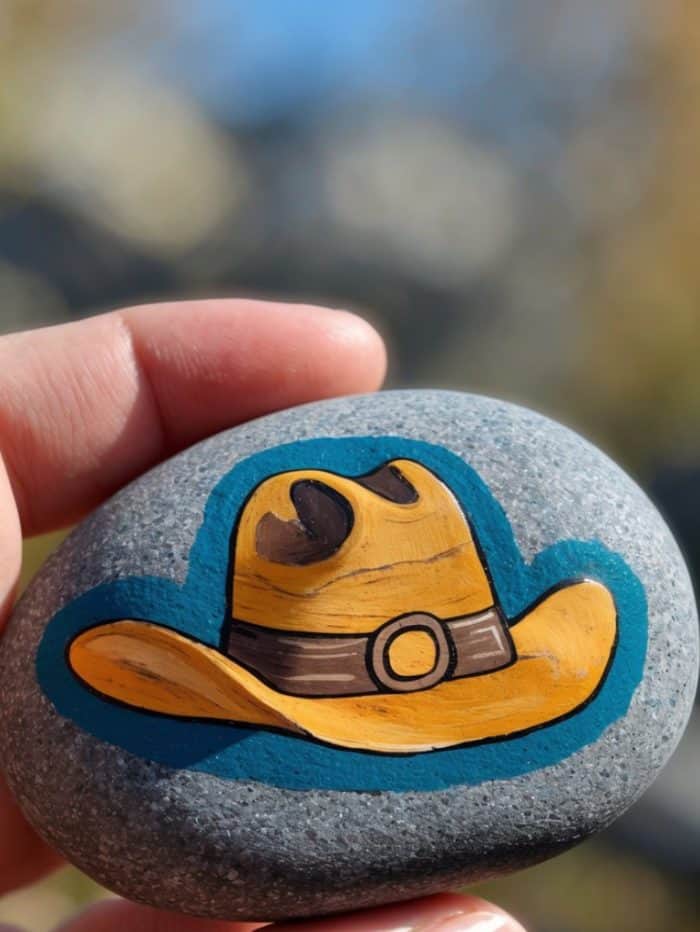 A hand holds a smooth gray stone painted with a yellow cowboy hat featuring a brown band and buckle, outlined in blue. The background is softly blurred.
