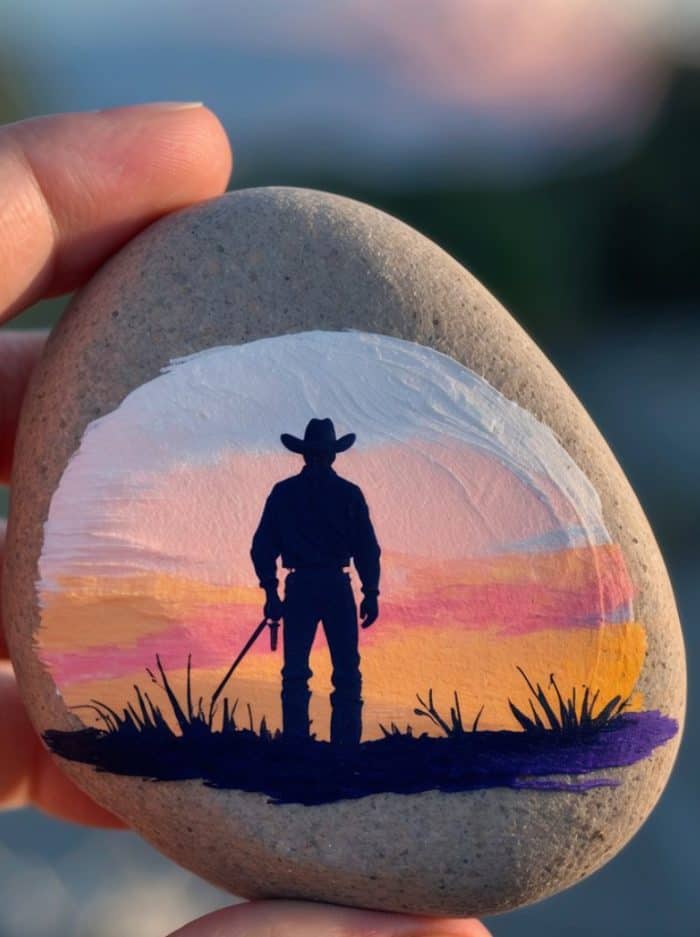 A hand holds a smooth stone painted with the silhouette of a cowboy holding a lasso, standing in grass against a colorful sunset sky in shades of pink, orange, and purple.