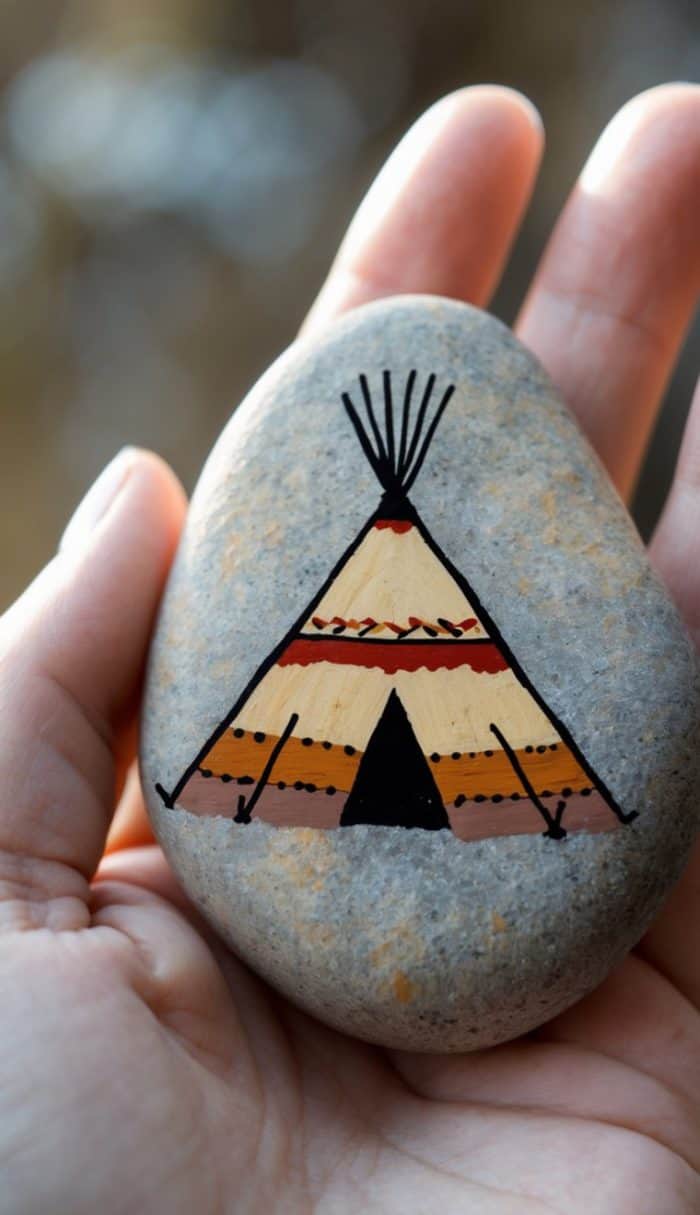 A hand holds a smooth stone painted with a colorful teepee design featuring geometric patterns in orange, red, black, and beige.