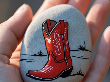 A hand holding a smooth stone painted with a red cowboy boot and simple black grass details on a light background.