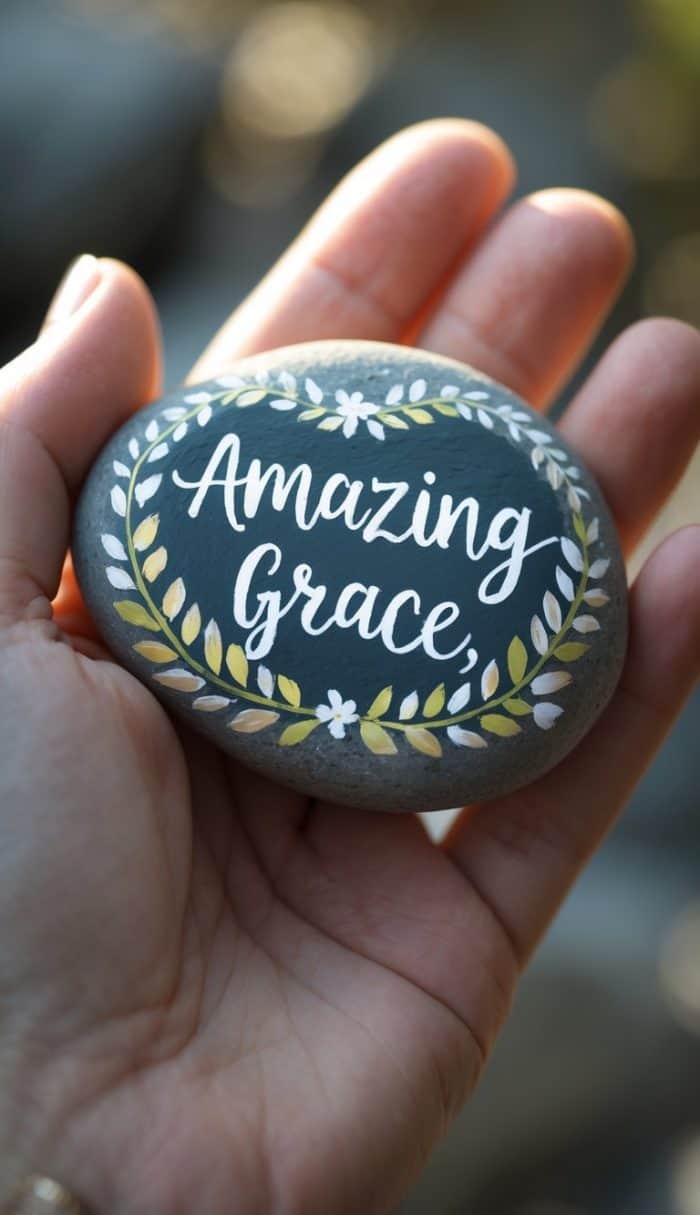 A hand holds a smooth stone painted with the words "Amazing Grace" surrounded by a decorative wreath of green and yellow leaves and small white flowers.