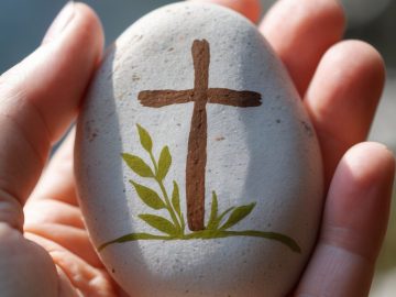A hand holds a smooth stone painted with a brown cross and green leaves, symbolizing Christianity and new life. The background is softly blurred.