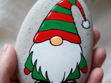 A hand holds a smooth stone painted with a festive gnome wearing a red and green striped hat, green clothes, and a white beard. A soft pine branch and white fabric are in the blurred background.