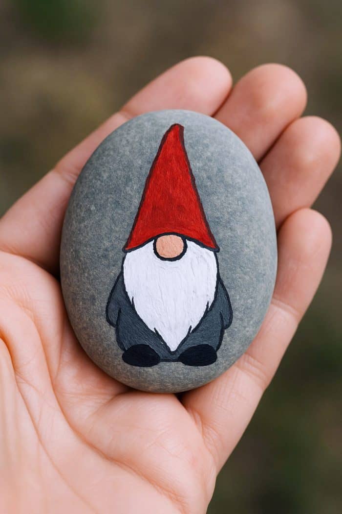 A hand holds a smooth gray stone painted with a gnome wearing a tall red hat, white beard, and dark clothing, against a blurred background.