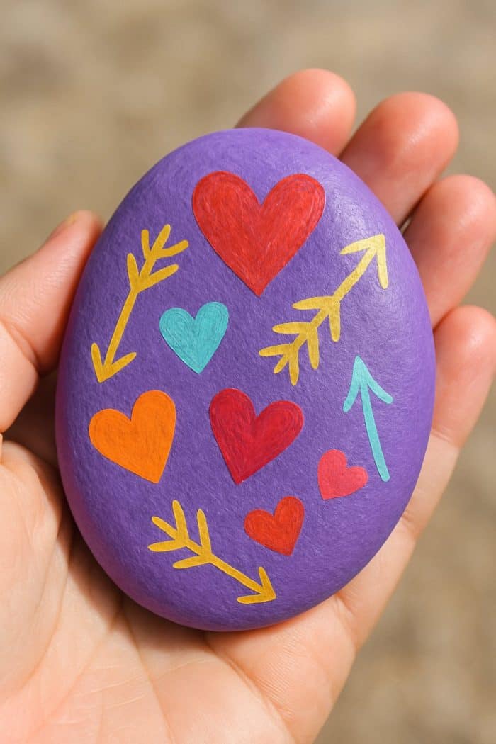 A hand holds a smooth, purple-painted stone decorated with colorful hearts and arrows in red, orange, teal, and yellow.