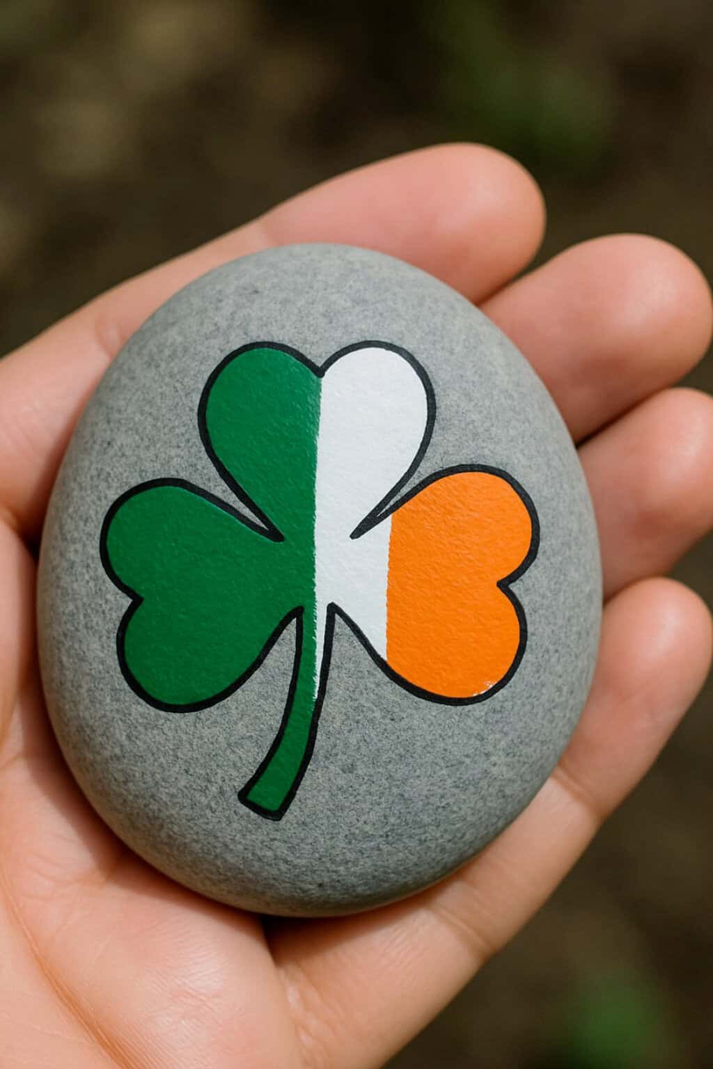 A hand holds a smooth gray stone painted with a shamrock in the colors of the Irish flag: green, white, and orange.