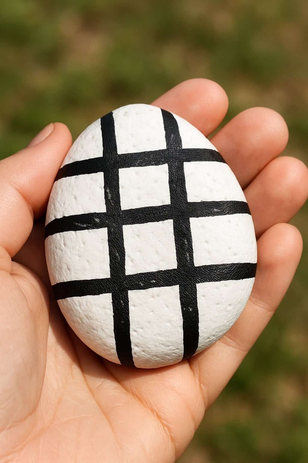 A hand holding a smooth white rock painted with black crosshatch patterns.