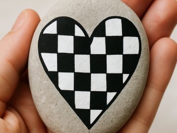A hand holds a smooth gray stone with a heart shape painted on it in a black and white checkerboard pattern.