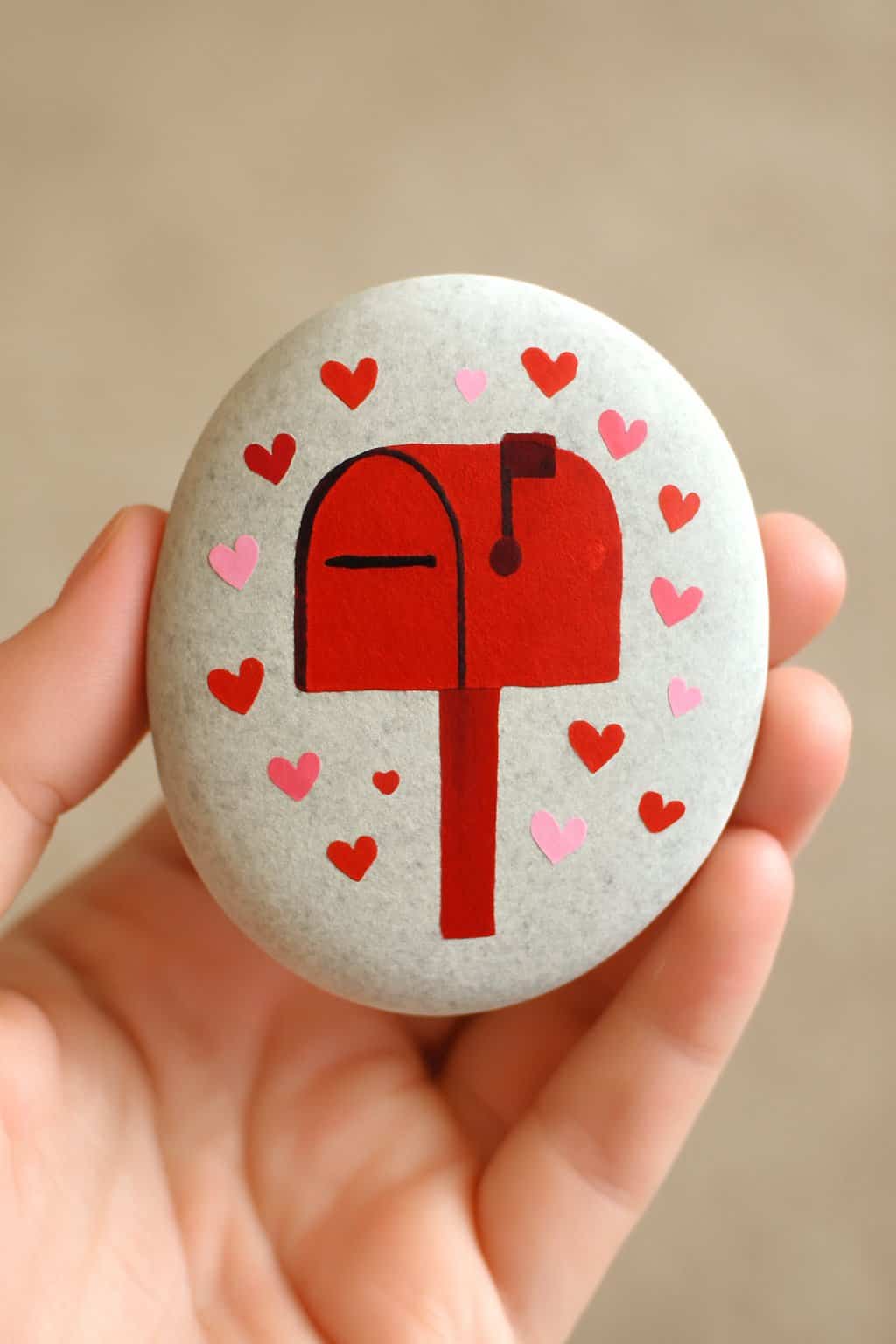 A hand holding a smooth river rock painted as a Valentine mailbox with hearts.