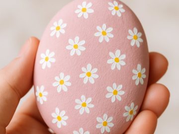 A hand holds a pink egg decorated with a pattern of small white and yellow daisy flowers. The background is softly blurred.