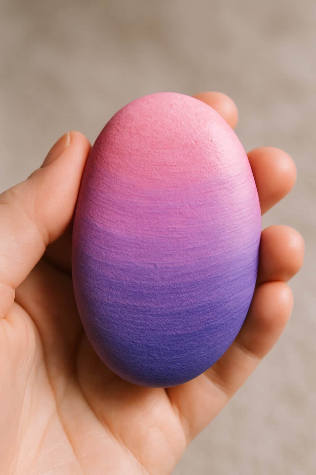 A hand holding a smooth river rock painted with pink and purple gradient colors.