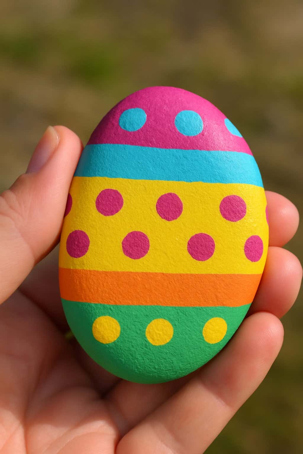 A hand holding a smooth river rock painted with colorful polka dots and stripes.
