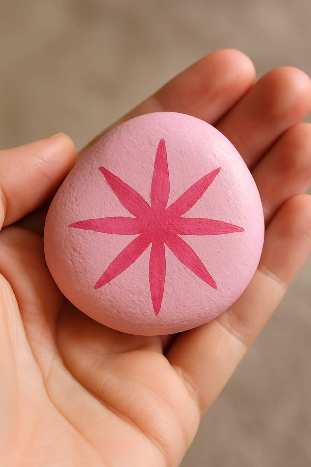 A hand holding a smooth river rock painted pink with a starburst design.