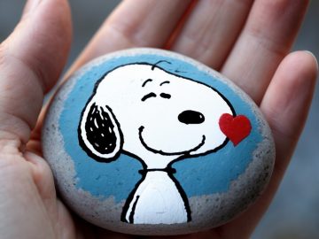 A hand holds a smooth stone painted with a smiling cartoon dog, resembling Snoopy, with a red heart near its cheek and a blue background.