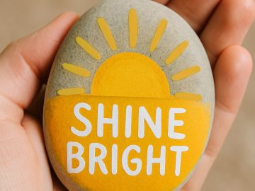 A hand holds a smooth stone painted with a yellow sun and the words "SHINE BRIGHT" in white letters.