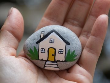 A hand holds a smooth, gray stone painted with a small house featuring a yellow door, black roof, windows, steps, and green bushes on either side.