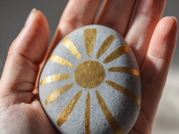A hand holds a smooth gray stone painted with a bright gold sunburst design, with sunlight softly illuminating the scene.