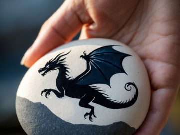 A hand holds a smooth stone painted with a black dragon featuring wings, standing on a gray, rocky background.