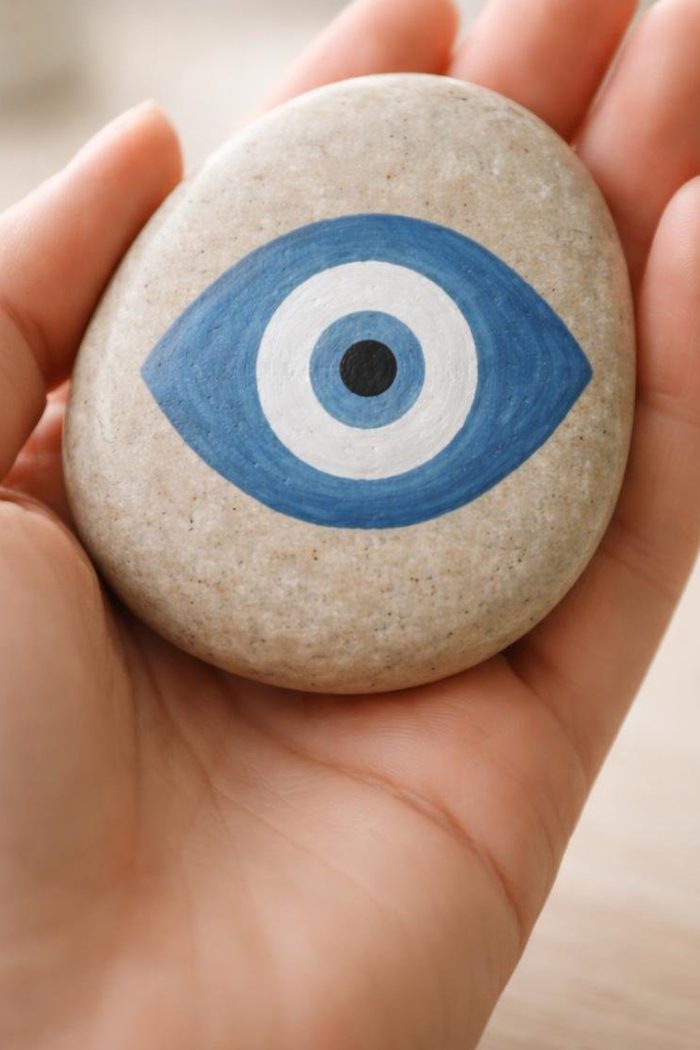 A hand holds a smooth stone painted with a blue and white eye design, resembling the evil eye symbol.