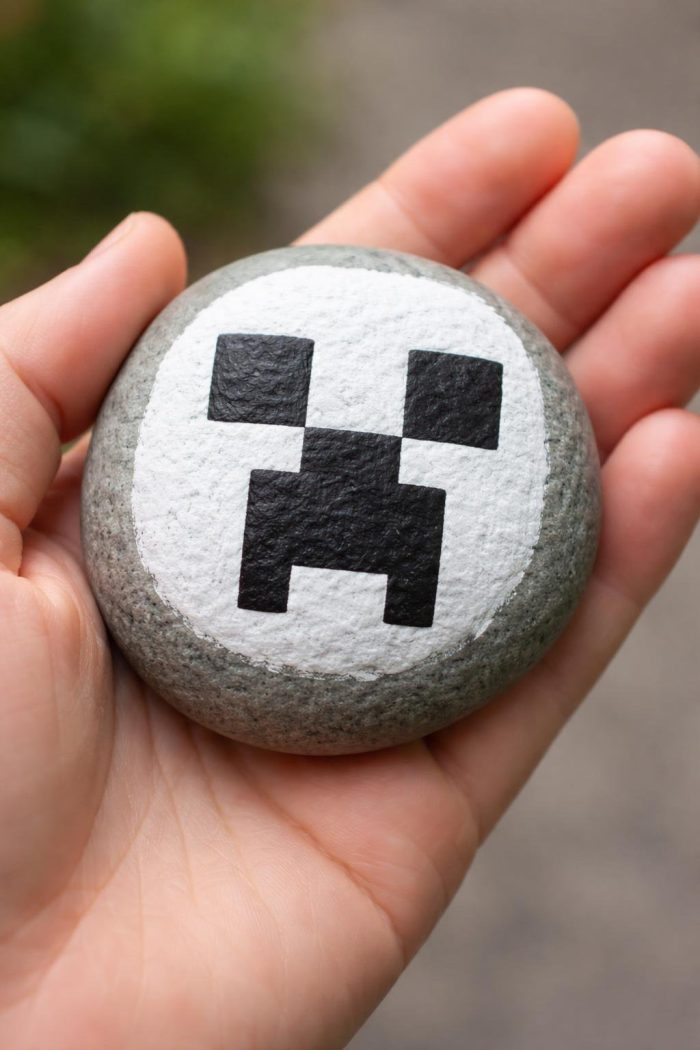 A hand holds a round stone painted with a white circle and a black pixelated face resembling the Creeper character from Minecraft.