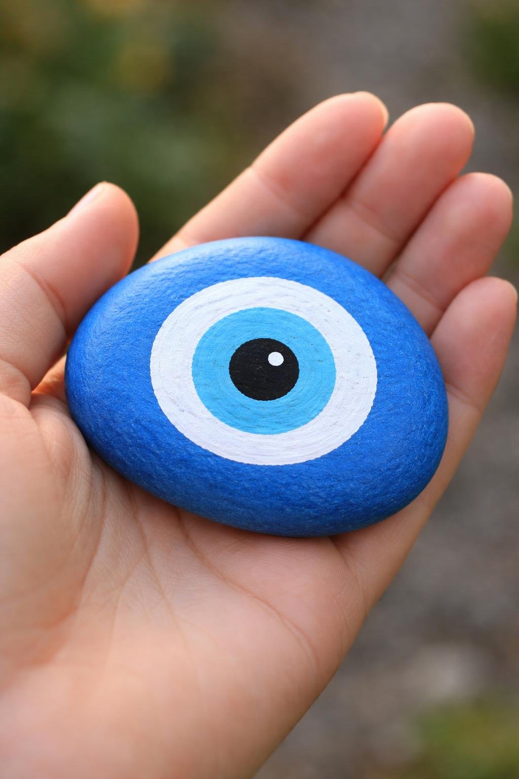 A hand holding a smooth river rock painted with a blue and white evil eye design.